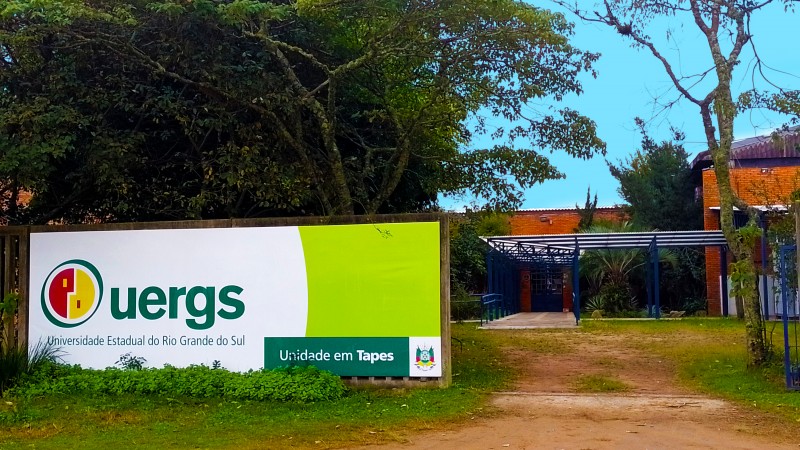 Em destaque, muro com a placa de identificação da unidade. Ao fundo, árvores e entrada de prédio.