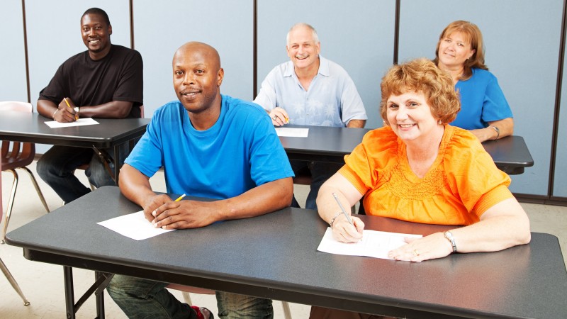 Uma sala de aula com 5 adultos: atrás, um negro de camiseta preta, um grisalho de camisa azul clara, uma loura de blusa azul; à frente, um negro de camiseta azul e uma senhora loura de blusa laranja, todos sentados em classes com papel e caneta.