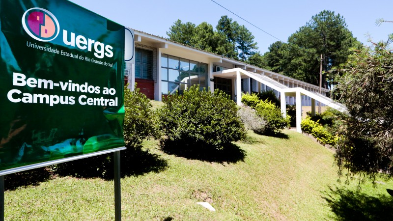 Fachada de um dos prédios do Campus Central, com grama e vegetação à frente. à esquerda, um banner com fundo verde e, em letras brancas, a frase "Bem-vindos ao Campus Central". Acima, o logotipo da Uergs.