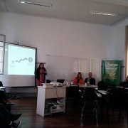 Pessoas reunidas em uma sala de aula, assistindo a uma mulher que faz uma apresentação com um telão.