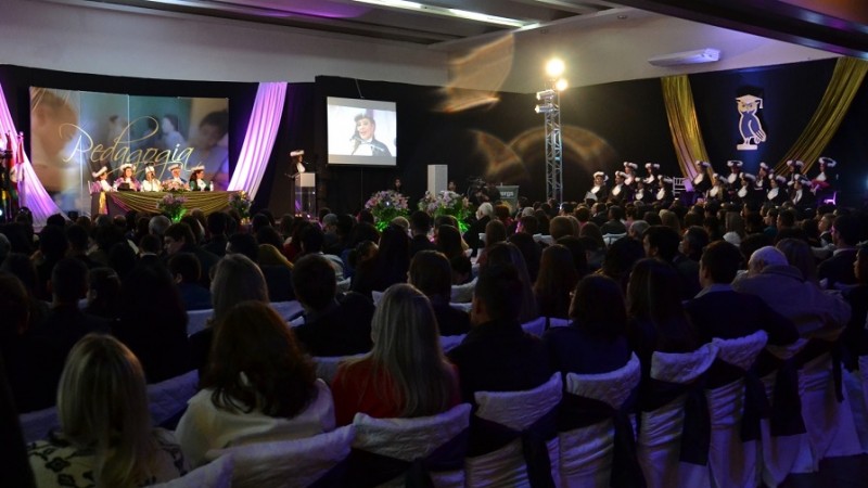 Formatura: público numeroso sentado e olhando para o palco, onde estão 5 autoridades acadêmicas em uma mesa, ao centro, e 16 formandos ao lado esquerdo da mesa. Ao fundo, imagens de criançaS e a palavra PEDAGOGIA.