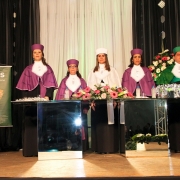 Mesa das autoridades acadêmicas, formada por cinco professores, que estão em pé. A reitora em exercício tem veste em branco, o paraninfo em verde e as pedagogas em roxo. Ao lado esquerdo da foto, banner da Uergs com mapa do Estado. 