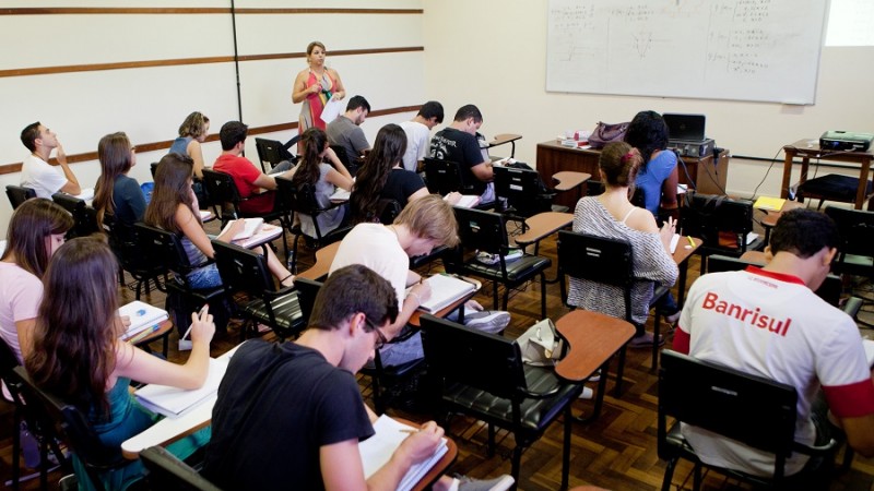 Sala de aula com estudantes sentados em suas mesas, de costas para o fotógrafo. À esquerda, uma professora em pé e, em frente aos estudantes, a mesa do professor e uma lousa branca.