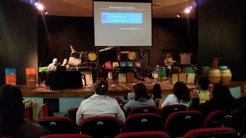 Público olhando em direção ao palco onde, à esquerda, estão o palestrante e a mediadora. Pendurado ao teto, um telão com as informações da palestra e, no palco, instrumentos de percussão para uma apresentação posterior.