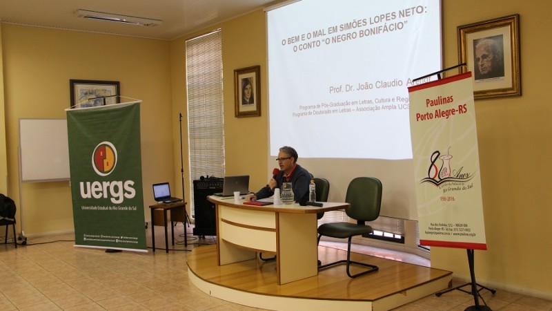 Palestrante sentado atrás de uma mesa. Acima, um painel projetando o tema da palestra. À esquerda, banner da Uergs e à direita banner com logotipo comemorativo pelos 80 anos da Livraria Paulinas.