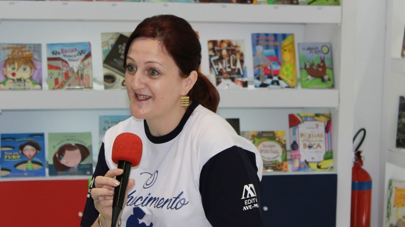 Enquadramento da foto em primeiro plano. Mulher de cabelos escuros e presos em rabo de cavalo, olhos verdes, com expressão alegre enquanto fala ao microfone, o qual segura com a mão direita. Atrás, prateleiras brancas com livros infantis.