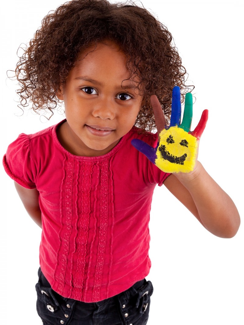 Menina negra vestida com blusa rosa e calça jeans, com o braço direito escondido atrás das costas e a mão esquerda levantada. Na palma da mão, a pintura de um rosto sobre um fundo amarelo e os dedos coloridos.