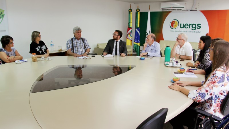 Pessoas sentadas ao redor de uma mesa bege com centro de vidro. Ao fundo, painel verde e laranja com logotipo da Uergs sobre fundo branco; à esquerda, bandeiras do Rio Grande do Sul, Nacional e da Uergs.