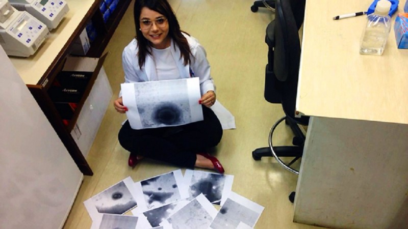 Jovem sorridente, com cabelos longos e escuros, usando óculos de grau, está sentada no chão e segura uma impressão com a imagem do vírus gigante. À sua frente e no chão, outras impressões. Ao seu redor, móveis em um laboratório.