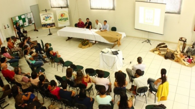 Plateia sentada em meio círculo, assistindo quatro expositores que estão sentados atrás de uma mesa. O ambiente é amplo e claro.