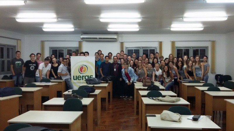 Sala de aula com mesas e cadeiras e, ao fundo, ao redor de um banner com o logotipo da Uergs, o grupo de participantes da aula inaugural.