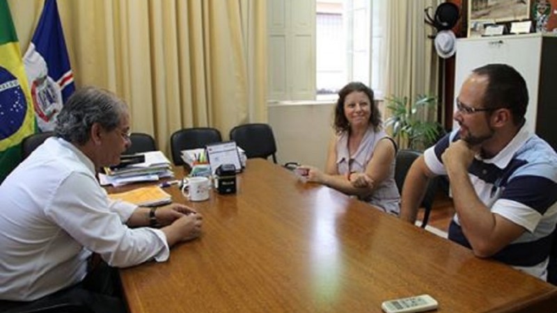 Ao redor de uma mesa, o prefeito com os professores Rodrigo e Carmen à sua frente. Ao fundo, uma janela com cortina e à esquerda, as bandeiras nacional e de Montenegro.