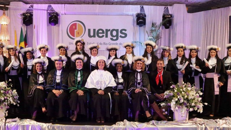 Sobre o palco e em pé, turma de formandos vestindo toga; em frente a eles e sentados, a reitora, paraninfos e homenageados. Ao fundo, painel com o logotipo da Uergs.