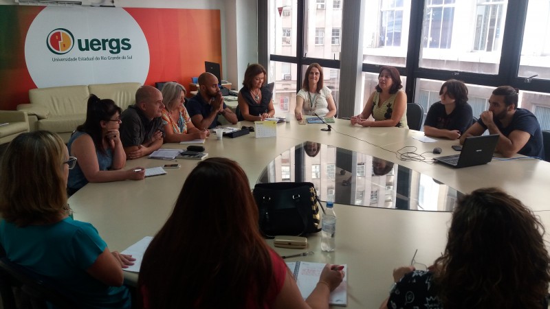 Ao redor de uma mesa, os participantes da reunião. Ao fundo da sala, um painel com o logotipo da Uergs.