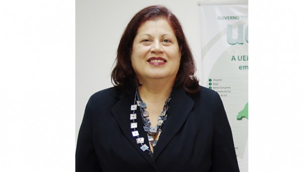 Mulher madura, de pele clara, com cabelos escuros até o ombro, sorridente, com batom em tom vermelho. Veste um blazer preto e usa um colar saliente, composto por peças quadradas furta-cor.