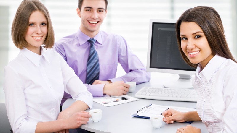 Duas mulheres e um homem sorridentes estão sentados ao redor de uma mesa, segurando uma xícara de café cada um. Ao fundo e sobre a mesa, um computador.