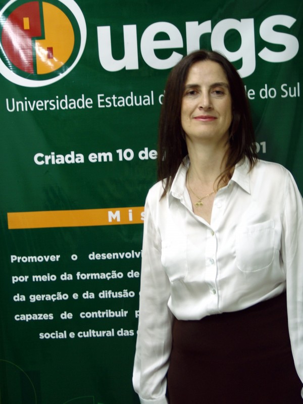 Mulher de meia-idade, cabelos escuros até o ombro, pele clara, sorriso discreto. Veste camisa de cetim branca e saia preta. Ao fundo, banner verde, com o logotipo e missão da Uergs em letras brancas.