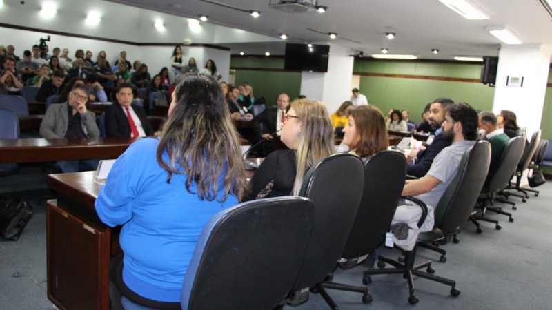 De costas para a foto, os integrantes da mesa sentados de frente para o público que ocupa todo o espaço.