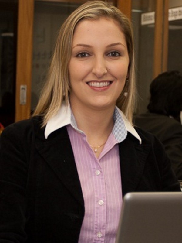 Mulher sorridente de pele cara, olhos castanhos e cabelo louro, liso e comprido. Veste camisa rosa e casaco preto.