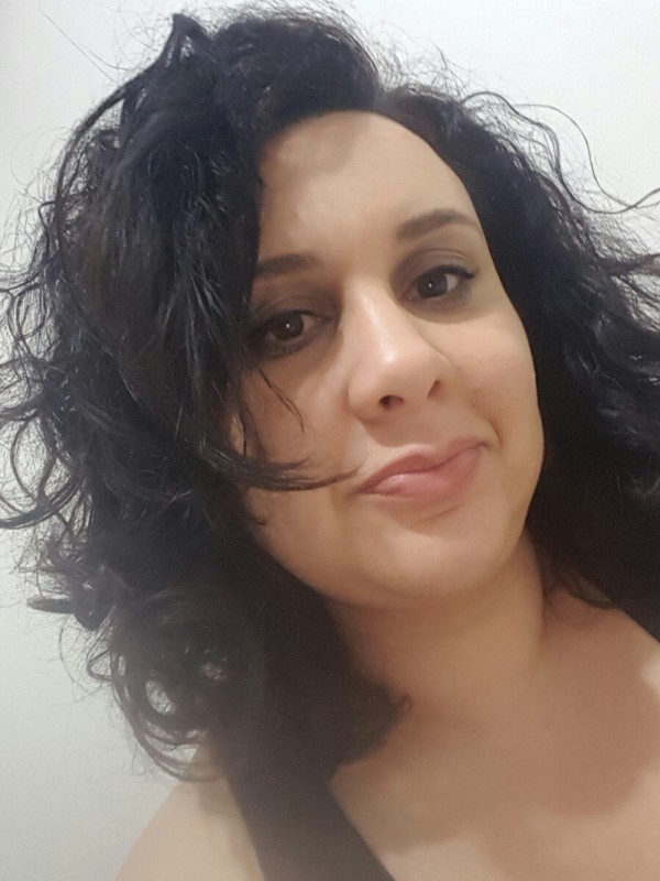 Mulher de pele cara, olhos castanhos, cabelo preto cacheado, na altura dos ombros. Veste regata preta.