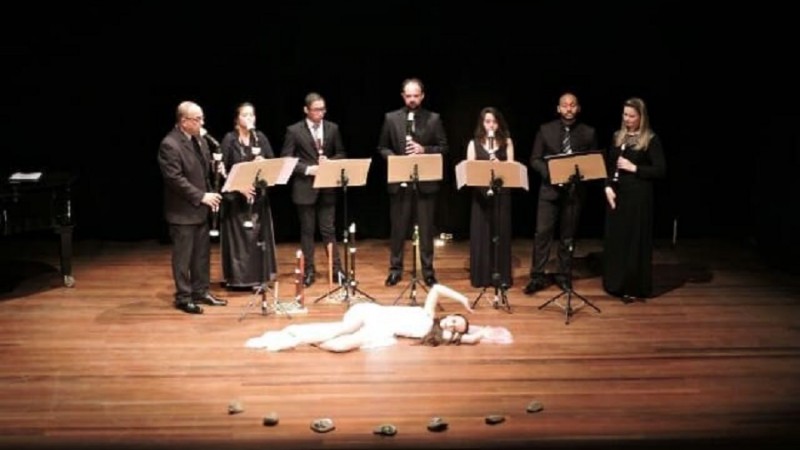 Em um palco, sete pessoas em pé, vestidas com traje social preto, cada uma com sua flauta doce, têm à frente cinco estantes com partituras. Deitada no palco, em frente às estantes, uma mulher vestida de branco parece realizar uma performance artística.