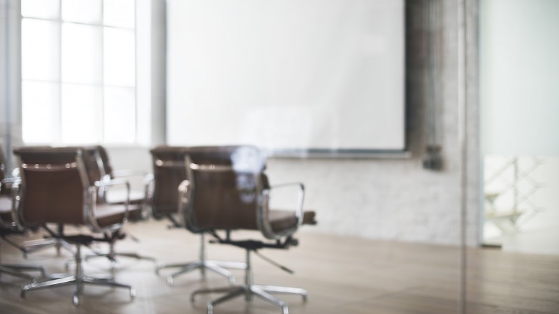 Sala de aula desfocada, com lona para projeção e 4 cadeiras giratórias marrons.