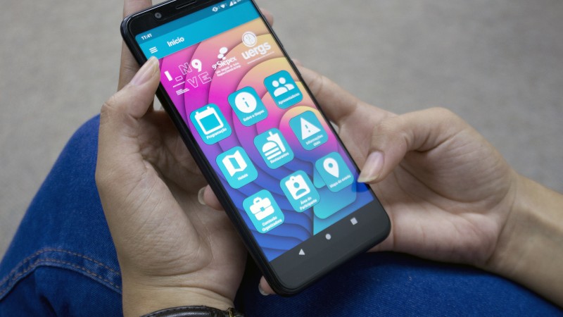 M&atilde;os femininas morenas seguram um smartphone. Na tela dele, a imagem da tela inicial do aplicativo do evento.