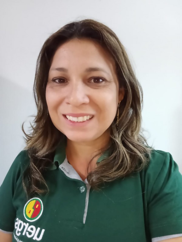 Mulher sorridente de pele clara, olhos castanhos, cabelos louro escuro com mechas, ondulados, na altura dos ombros. Veste camisa polo verde com o logotipo da Uergs.