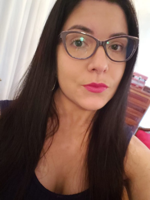 Mulher de pele clara, cabelos escuros, lisos e compridos. Usa óculos de grau cinza e batom rosa.