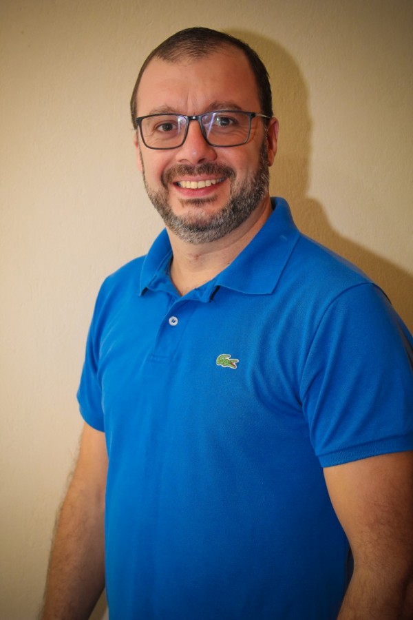 Homem sorridente de pele clara, cabelo castanho, barba e bigode grisalhos. Usa óculos de grau e veste camisa polo azul.