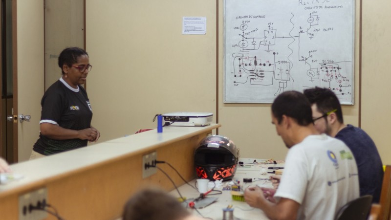 Em uma sala de aula, dois alunos sentados realizam atividades. Uma professora se aproxima de onde eles estão.