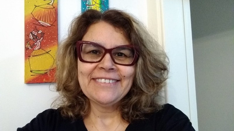 Mulher de meia-idade, branca, de cabelos loiros que vão até os ombros, sorridente. Ela usa óculos de grau na cor bordô e camiseta preta. 