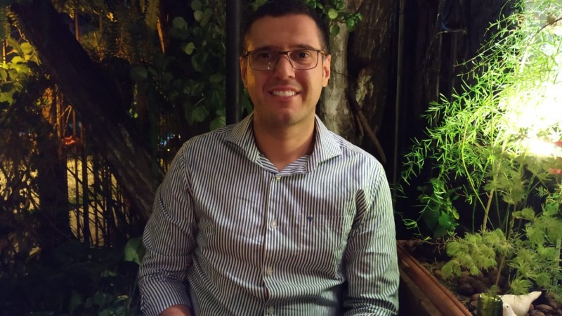 Homem de pele clara, olhos e cabelos castanhos escuros. Está sorridente. Usa óculos de grau e camisa listrada em azul e branco. Ao seu redor, árvores e folhagens.