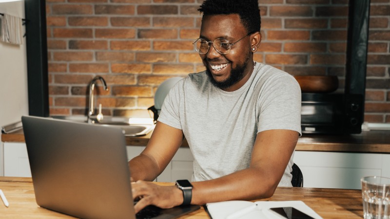 Foto colorida de um jovem negro sorridente em frente a um notebook, com uma parede de tijolos aparentes e uma pia de cozinha ao fundo.