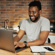 Foto colorida de um jovem negro sorridente em frente a um notebook, com uma parede de tijolos aparentes e uma pia de cozinha ao fundo.