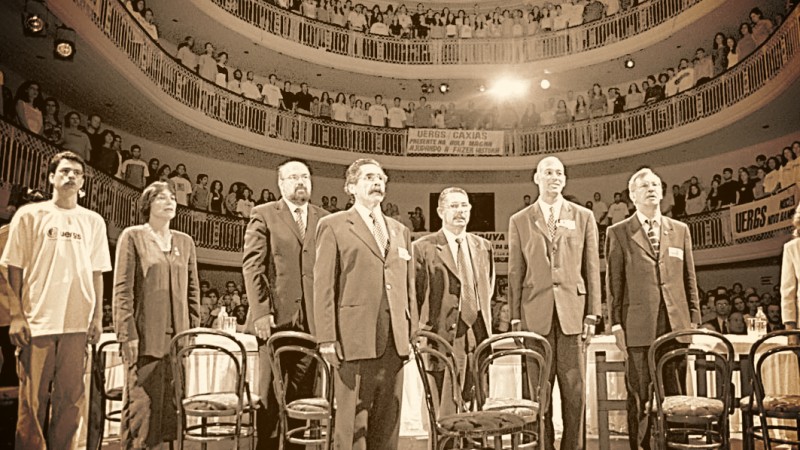 Ao centro, foto em tom sépia da primeira Aula Magna da Uergs, no dia 8 de abril de 2002. No retrato, há sete pessoas em postura ereta, com os braços distendidos ao longo do corpo, o que sugere a execução de um hino. Dentre elas, o governador Olívio Dutra.