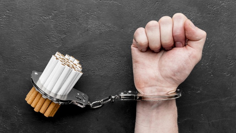 Sobre fundo cinza escuro, algema prendendo um maço de cigarros e o punho direito de uma pessoa branca.