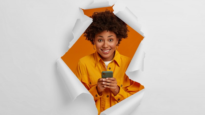 Imagem com fundo claro, imitando um papel rasgado no centro, revelando a imagem de uma jovem negra de jaqueta laranja, sobre um fundo laranja, segurando um smartphone com as duas mãos e sorrindo com expressão de surpresa.