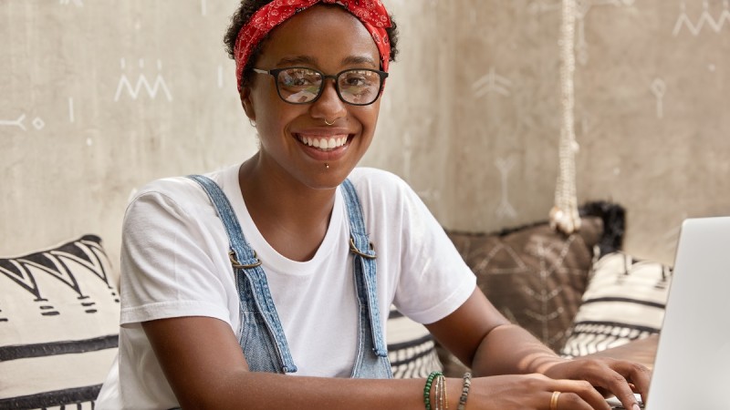 Foto colorida. Jovem negra sorridente, olha para a câmera enqaunto digita em um noteebook. Ela usa óculos de grau, faixa vermleha nos cabelos curtos, blusa branca e macacão jeans.