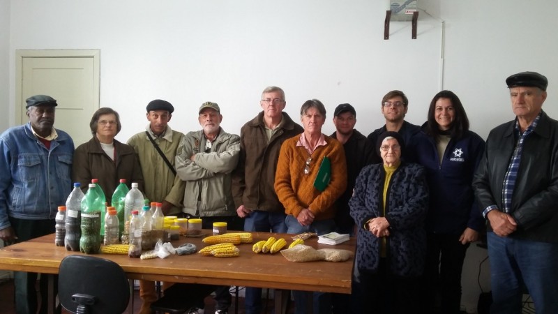 Ao redor de uma mesa com sementes em garrafas plásticas e espigas de milhos, estão onze pessoas em pé.