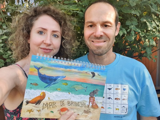 Foto colorida: mulher branca de olhos azuis e homem branco de camiseta azul posam sorridentes segurando um livro com ilustração de mar com animais marinhos.