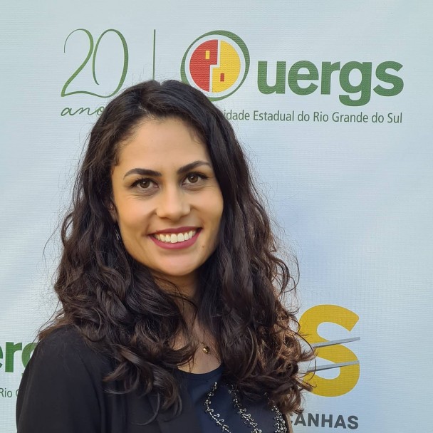 Foto colorida. Mulher morena clara, sorridente, de cabelos ondulados longos e olhos castanhos escuros. Acima de sua cabeça, o selo de 20 anos da Uergs.