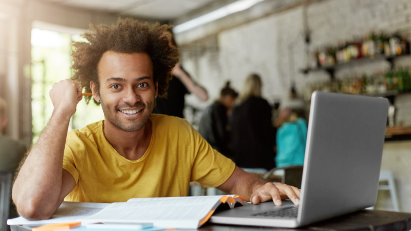 Um jovem negro de pele clara, com cabelo crespo volumoso, sorridente, sentado com livros e um notebook à sua frente. 