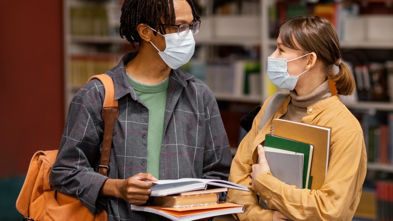 Dois estudantes na biblioteca, com livros na mão e usando máscara de proteção facial.