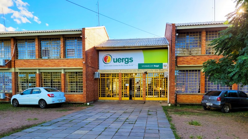 Foto colorida de um prédio horizontal de dois andares, com paredes de tijolos à vista, janelas e portas de vidro. Ao centro, sobre a porta de entrada, uma placa nas cores branca e verde limão com o logotipo da Uergs e o texto Unidade em Bagé.