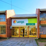 Foto colorida de um prédio horizontal de dois andares, com paredes de tijolos à vista, janelas e portas de vidro. Ao centro, sobre a porta de entrada, uma placa nas cores branca e verde limão com o logotipo da Uergs e o texto Unidade em Bagé.