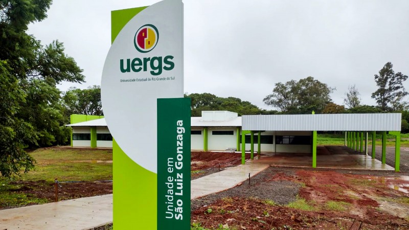 Foto colorida de um terreno com grama e terra vermelha, com um pavilhão ao fundo, com paredes brancas e vigas em verde limão. Em primeiro plano, um totem nas cores branca, verde limão e verde escuro, com o logotipo da Uergs e o texto Unidade em São Luiz Gonzaga.