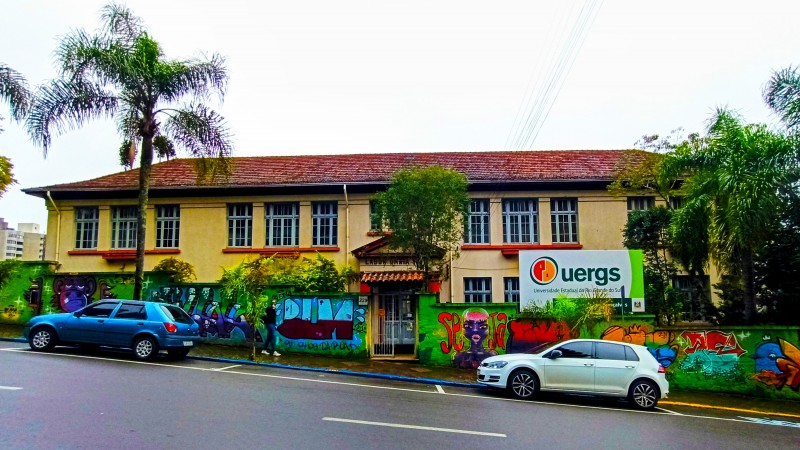 À frente, muro com muitos desenhos coloridos e a placa de identificação da Uergs acima dele. Ao fundo, prédio de 2 andares com paredes em tom de bege.