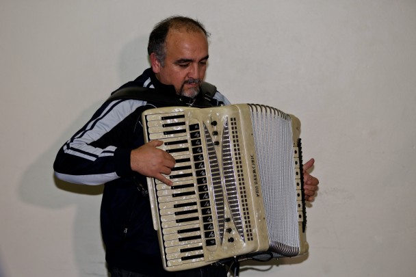 Homem de pele morena clara, cabelos ralos grisalhos, com barba grisalha. Toca um acordeon de cor clara perolada.