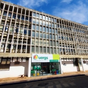 Foto de um prédio grande com muitas janelas envidraçadas. Ao centro, sobre a porta de entrada, a placa de identificação da Uergs.